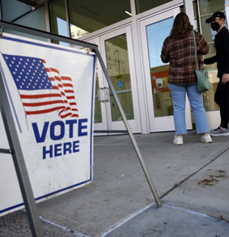 2 voters at their polling site next to a sign that reads VOTE HERE