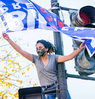 Woman wearing a face mask while standing on a light post holding up a Biden for President 2020 flag 